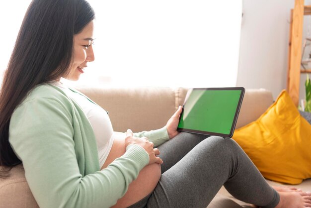 pregnant woman hands fondle on belly and holding green chroma key screen on tablet sitting on sofa