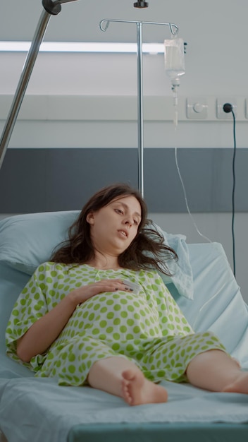 病棟のベッドに横になり、出産の支援を待っている妊婦。クリニックで一緒に座って出産する子供を期待している妻のためのバックパックを保持している白人男性