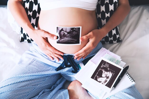 Pregnant woman makes notes in notebook and holding ultrasound image and medical document
