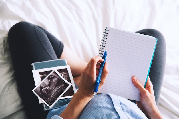 Pregnant woman makes notes in notebook and holding ultrasound image and medical documents