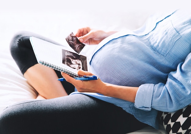 Pregnant woman makes notes in notebook and holding ultrasound image and medical documents