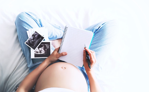 Pregnant woman makes notes in notebook and holding ultrasound image and medical documents