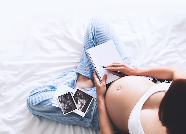 Pregnant woman makes notes in notebook and holding ultrasound image and medical documents