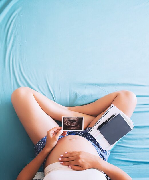Pregnant woman makes notes in notebook and holding ultrasound image and medical documents