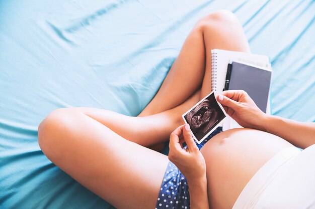 Pregnant woman makes notes in notebook and holding ultrasound image and medical documents