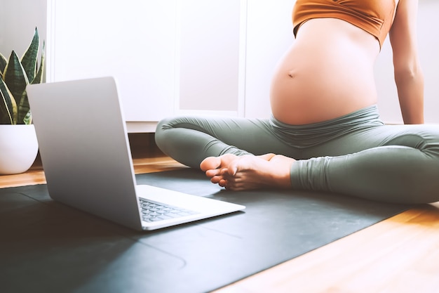 Беременная женщина занимается йогой дома с ноутбуком. женские упражнения медитируют во время беременности.