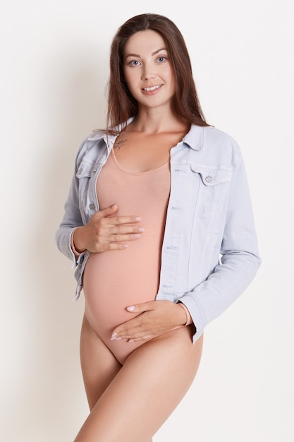 Pregnant woman in rosy bodysuit and denim jacket posing