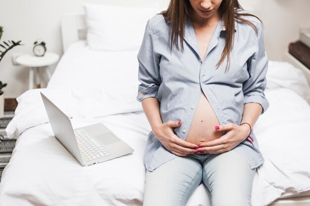 A pregnant woman sitting on bed near the laptop holding her belly with two hands