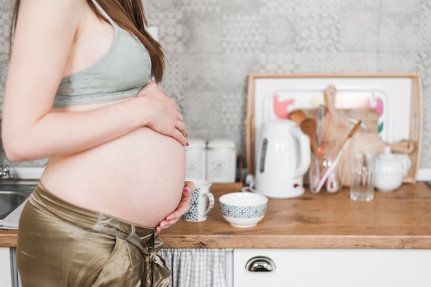 Беременная женщина, стоящая рядом с кухонным прилавком, касаясь ее живота