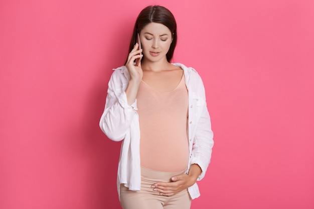 임신 한 여자는 그녀의 스마트 폰으로 이야기하고 만지고, 내려다보고, 흰 셔츠를 입고