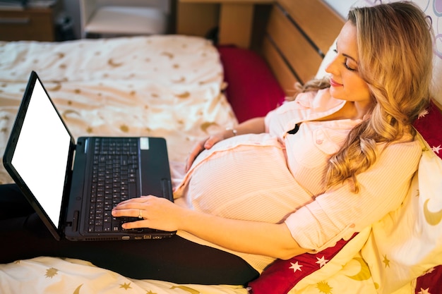 Donna incinta che per mezzo del computer portatile con lo schermo bianco in bianco