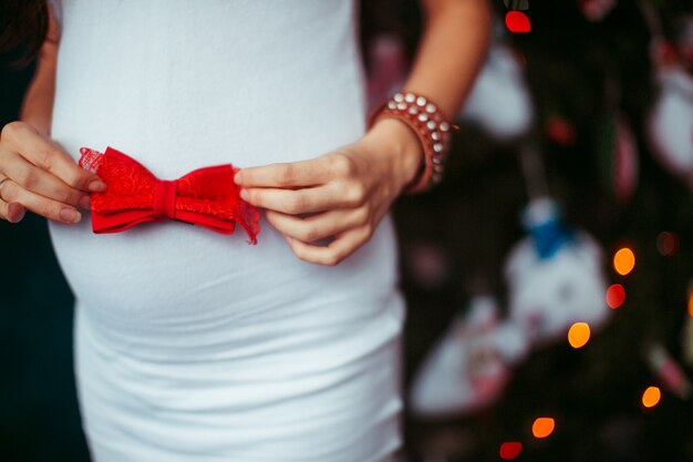 Pregnant woman in white dress holds red bow on her belly