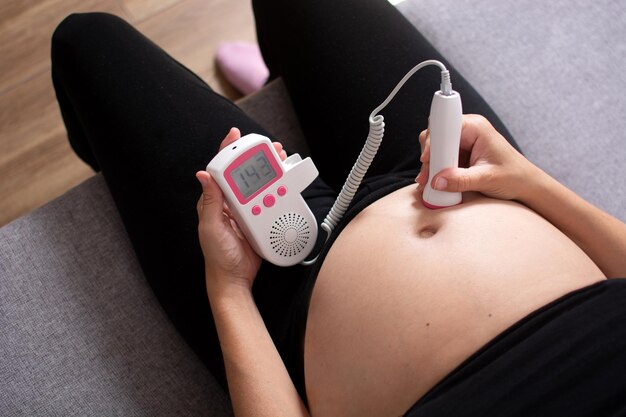 Pregnant woman with a fetal doppler listening to the baby's heart sitting on the sofa at home