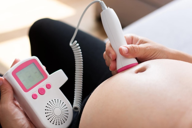 Беременная женщина с фетальным допплером слушает сердце ребенка, сидя дома на диване