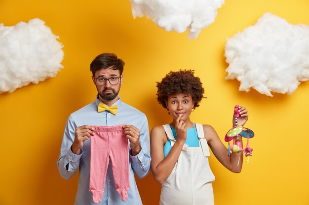 Pregnant woman with her husband prepare for child birth, buy clothes and toys for newborn, attend training courses for future parents, stand against yellow . Parenting and pregnancy concept