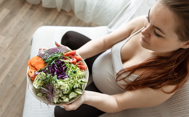 Foto gratuita donna incinta con un piatto di insalata di verdure fresche a casa sul divano vista dall'alto.