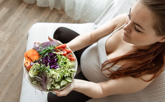 Pregnant woman with a plate of fresh vegetable salad at home on the couch top view.