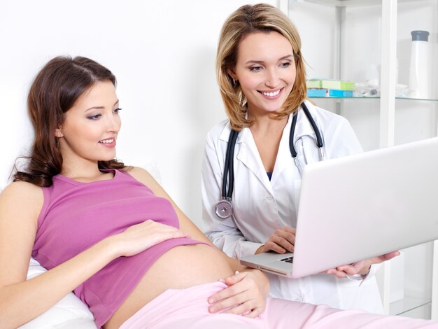 Беременная молодая женщина и ее врач, глядя на ноутбук - в помещении