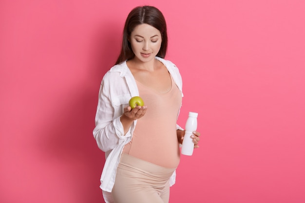 Pregnant young woman holding green apple and bottle of yogurt or milk.asual clothing.