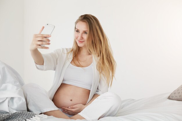 태아기 셀카. 그녀의 배꼽을 잡고 웃는 스마트 폰을 사용하여 자기 초상화를 만드는 임신 미래 엄마