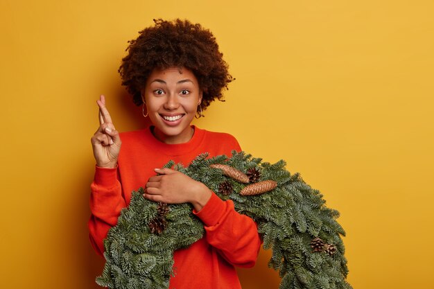 冬休みやお祭りのインテリアの準備。陽気なアフリカ系アメリカ人の女性が指を交差させ、願い事をし、クリスマスリースでポーズをとる