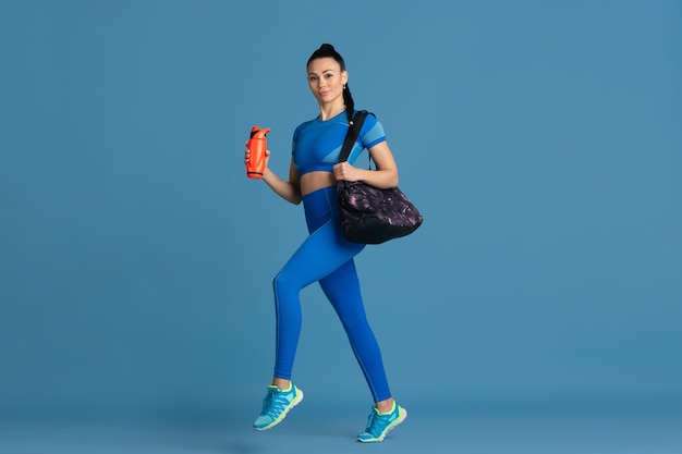 준비되었습니다. 아름 다운 젊은 여성 운동 선수 연습, 단색 파란색 초상화. 물병과 가방이 달린 스포티 한 맞는 갈색 머리 모델. 웰빙, 건강한 라이프 스타일, 아름다움 및 행동 개념.