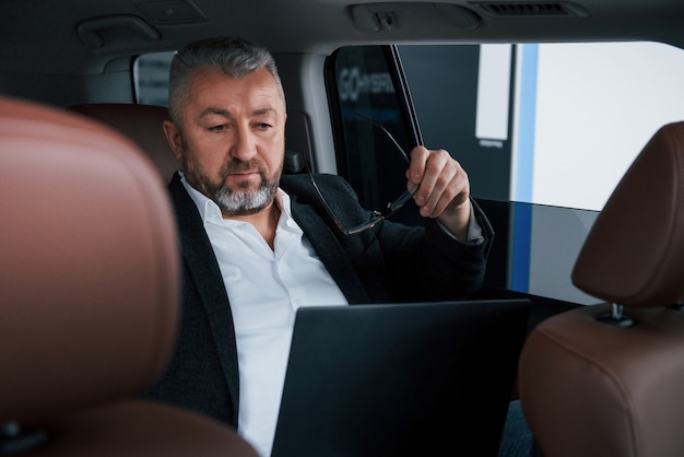 Preparing to read some documents. Working on a back of car using silver colored laptop. Senior businessman