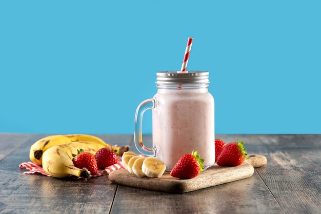 Preparing strawberry and banana smoothie in jar on wooden table and blue background