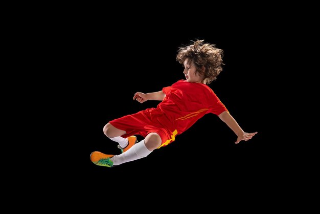 Foto gratuita giocatore di calcio in età prescolare in uniforme sportiva rossa che pratica con pallone da calcio isolato su sfondo scuro dello studio concetto di hobby del gioco sportivo