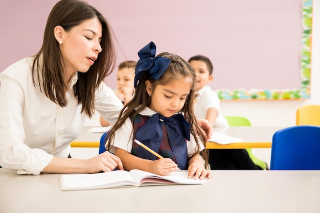 Preschool teacher sitting next to one of her students and helping her with her writing assignment