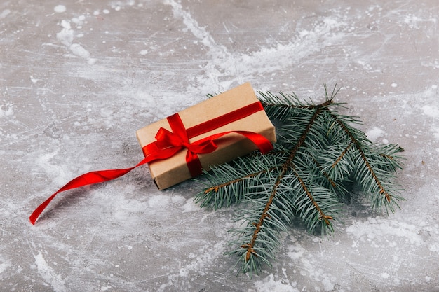 Present box made of grey brown paper and twined with red ribbon lies with a fir branch on grey floor