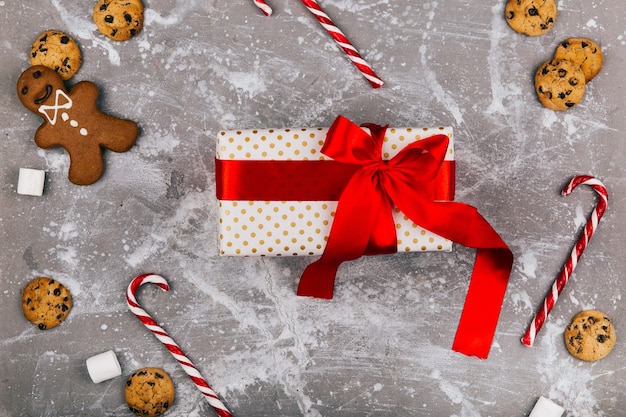 Present box with red ribbon lies on grey floor with Christmas cookies, gingerbreads and red white candies