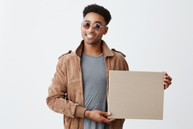 Present for you. Isolated on white portrait of young fashionable dark-skinned man with afro hairstyle in grey t-shirt, brown jacket and sun-glasses holding box in hand, smiling in camera.