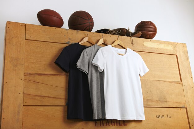 Презентация белых, серых и черных однотонных футболок с короткими рукавами, винтажных футбольных, баскетбольных и волейбольных и старых спортивных кожаных ботинок на деревянной транспортировочной коробке.