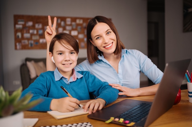 A Preteen boy uses a laptop to make a video call with his teacher next to his mother