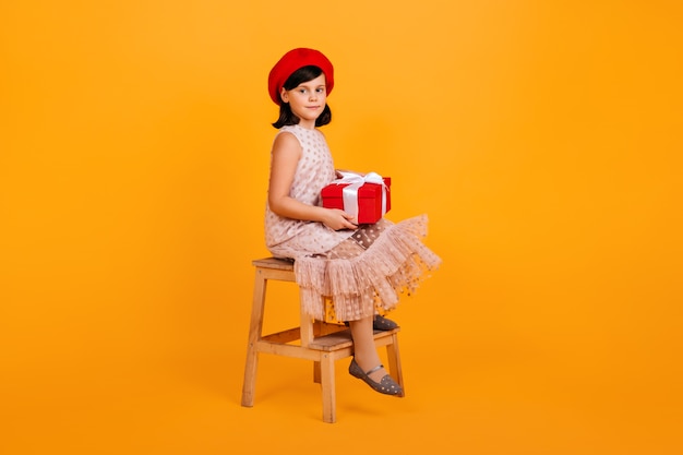 Ragazza del preteen in vestito che tiene regalo di compleanno. ragazzo con presente seduto su una sedia sul muro giallo.