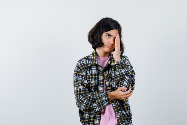 Preteen girl holding hand on eye in shirt,jacket and looking bored. front view.