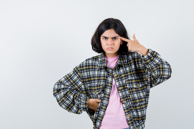 Девочка десятилетнего возраста в рубашке, куртке, указывающей на ее висок и сердитой, вид спереди.