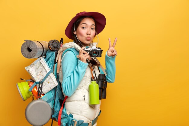 Pretty active backpacker makes victory gesture, keeps lips rounded, holds retro camera, stands with travelbag, takes photos during trip, isolated over yellow background