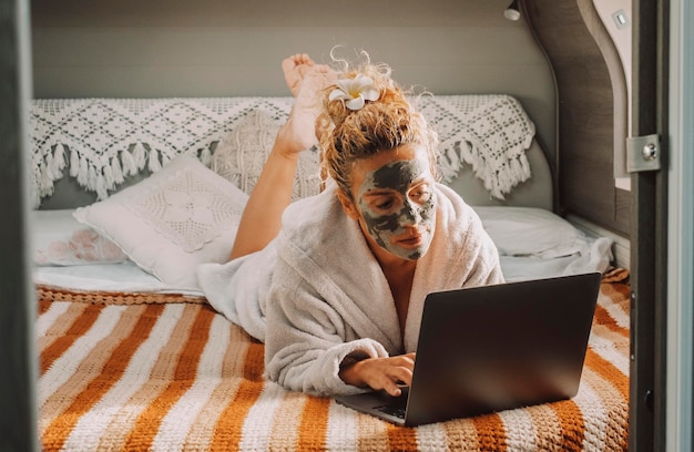 キャンピングカーの寝室に横たわっているラップトップコンピューターで顔に肌の美容クリームを塗ったかなり大人の女性。オンラインの仕事でライフスタイルの女性を旅行します。現代の女性と健康的な治療 Premium写真