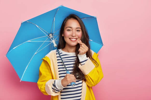 Pretty affectionate Asian woman makes korean like sign, has happy expression, gentle smile, stands under umbrella, wears yellow raincoat