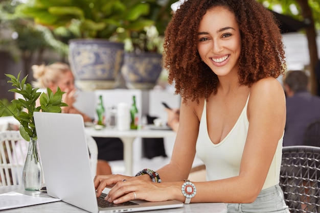 Free photo pretty african american female model keyboards something on laptop computer, connected to free wireless internet in cafe, writes new article for her blog