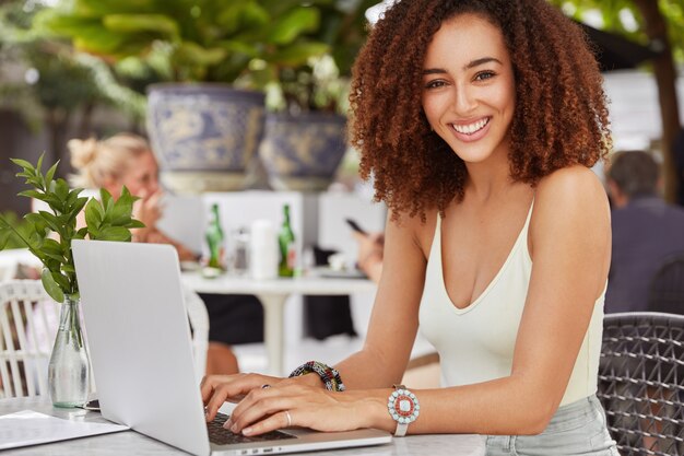 Pretty African American female model keyboards something on laptop computer, connected to free wireless internet in cafe, writes new article for her blog