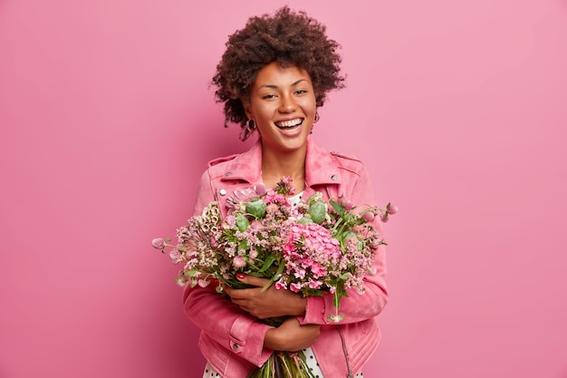 La bella donna afroamericana esprime emozioni sincere, abbraccia bouquet di fiori, ha umore primaverile