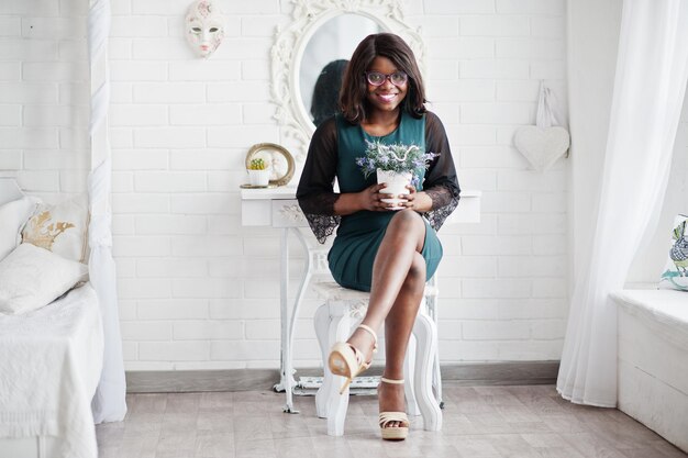 Симпатичная африканская американка в очках позирует в комнате у зеркала с цветочным горшком в руке