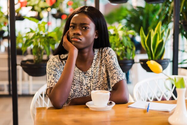 Pretty African-American woman sitting at table near laptop and looking with tired face expression.