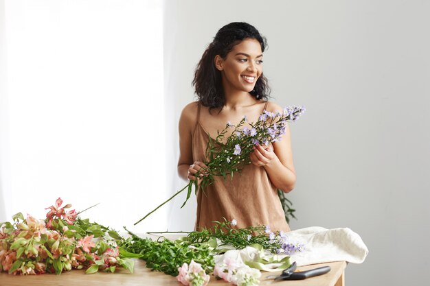 Pretty african female florist smiling making bouquet at workplace over white wall.