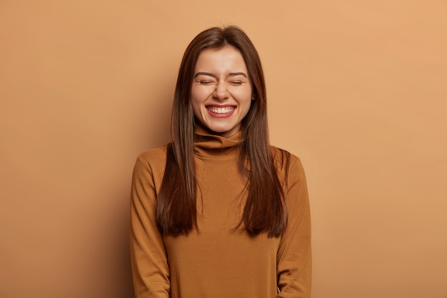 Pretty amused woman has long dark straight hair, laughs from joy, wears brown neck sweater, smirks face with pleasure