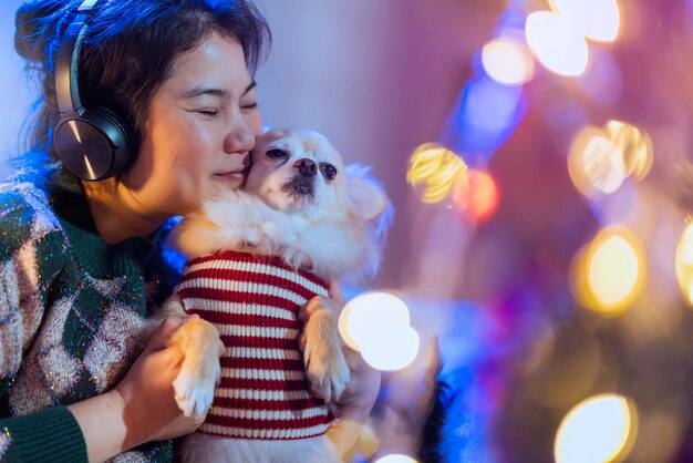 かなりアジアの女性は、注意と愛の優しさでかわいい小さなラップドッグの友人を抱きしめるヘッドフォンの手から音楽を聴きます陽気な瞬間アジアの女性は、クリスマスを飾る木とソファに小さなペットを抱きます