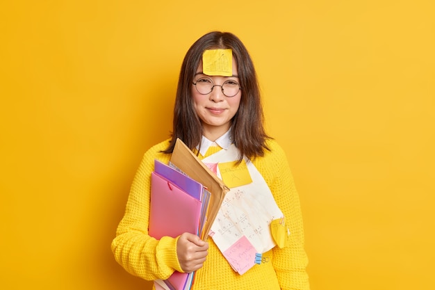 額にリマインダーステッカー紙を持ったかなりアジアの女子学生は、紙の入ったフォルダーを運び、難しいテストウェアの丸い眼鏡とジャンパーの準備をします。